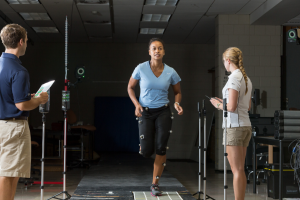 A student in a blue shirt is running on a sensor pad with sensors attached to her legs. Another student is standing nearby taking notes on a clipboard.