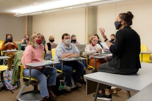 A masked professor sits on a table in front of a classroom of masked students. The professor is gesticulating.