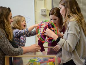 Three students are supervising a young child playing with a brightly colored toy that is being pulled out of a box.