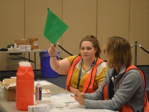 A student in an orange vest has a green flag raised signaling that there is room at her table to provide an individual with a flu shot.