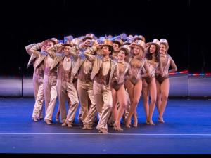 A group of actors in A Chorus Line dressed in gold tuxedos, leotards, and top hats form a triangle. All the actor hold the brim of their hat with their right hand and have their left leg crossed over the right.