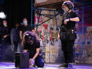 A Stage Management student dressed in all black stands next to another Stage Management student crouching down. The crouching student is taping a mark on the set of RENT.