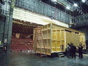 A large rectangle made of wood onstage is being constructed. A group of students stand by the set and a ladder is open next to the set.