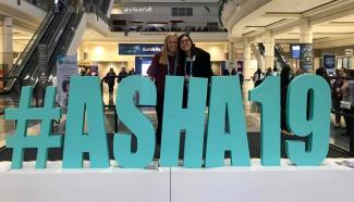 There are two students standing behind a large turquoise sign that reads #ASHA19