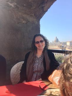 Woman in sunglasses and overcoat seated at cafe table with view of Saint Peters and Rome in distance.