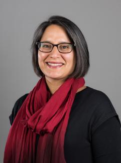 Headshot image of Belisa González. Belisa is framed by a grey background. Belisa is wearing a black long-sleeve top with a red scarf and glasses. Belisa's has darker hair that frames her face extending just above her shoulders. Belisa is looking directly at the camera and smiling.