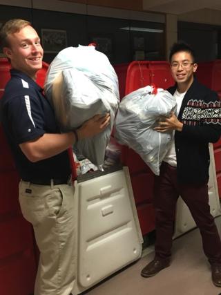 Two students in blue shirts and tan pants are holding large bags of clothes and getting ready to donate them.