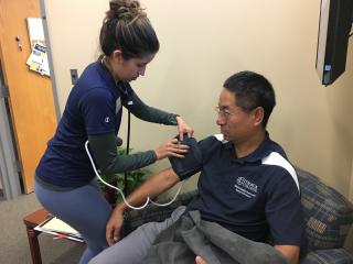 A student in a blue shirt is placing a blood pressure cuff on a man seated in a chair to take his blood pressure.