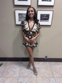 Desoni is posed for a photo in front of a beige wall with framed images. Desoni is wearing a short dress that is grey and black patterned and is smiling at the camera.