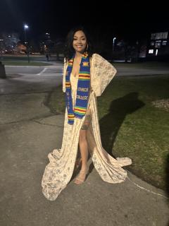 Heaven is posed for a photo at night. Heaven is wearing a long white patterned dress with a graduation stole. Heaven is facing the camera and smiling.