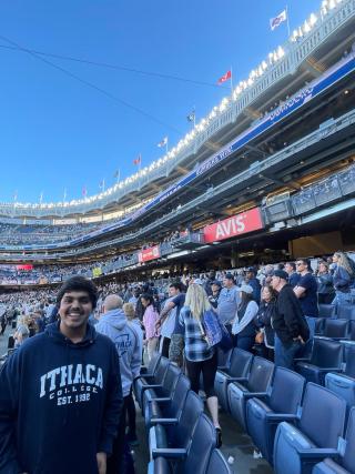 Utkarsh is inside Yankee Stadium for Cortaca. He is wearing a blue Ithaca hoodie with a crowd in the background. He is smiling for the camera.