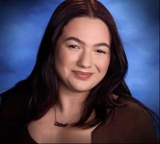 Viviana is posing for headshot. Is wearing a burgundy top and necklace. She has long hair. There is a blue background.