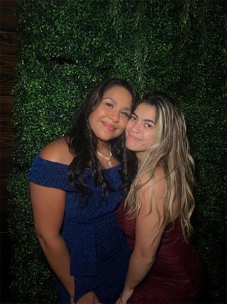 Alexa is posing with another individual in front of green textured background. Alexa is wearing blue dress and is smiling for the camera. Alexa is the individual on the left.