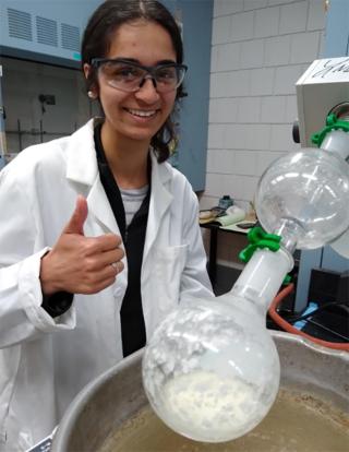 Ranjini is posing for the photo wearing a white coat and safety glasses in a lab. She has her right thumb up and is next to a instrument.