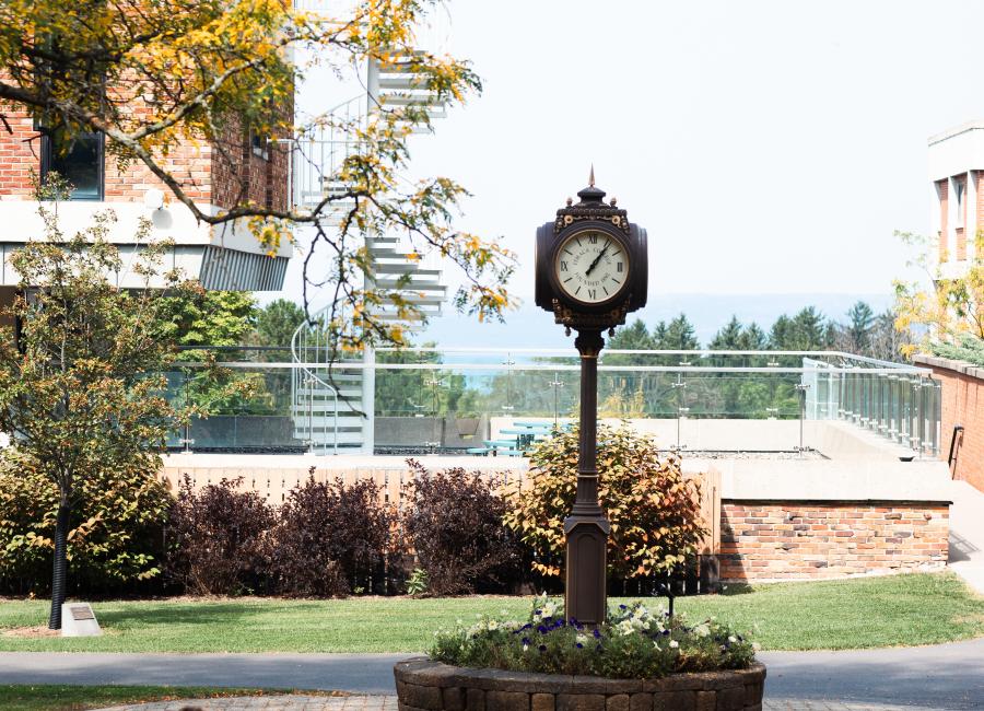 Image of Clock on Academic Quad. Student is sitting in foreground facing away from camera towards the clock. Leaves are scattered around the ground around the clock.