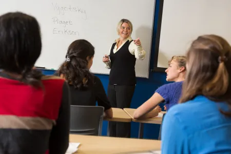 A faculty member in a blue vest and white shirt is standing at the front of a classroom, and has written Vygotsky, Piaget, and Eriksen on the white board. Four students, with their back turned toward the camera, are listening to the faculty member talk.