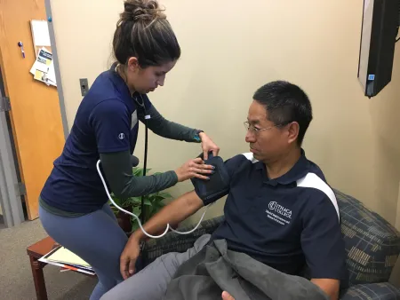 A student in a blue shirt is placing a blood pressure cuff on a man seated in a chair to take his blood pressure.