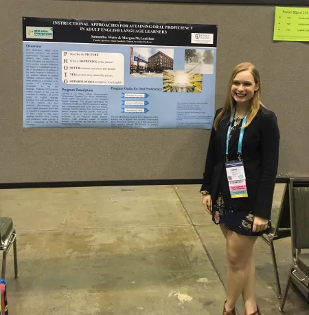 A student dressed in a blue suit is standing in front of a poster presentation on "Instructional Approaches for Attaining Oral Proficiency in Adult English Language Learners"