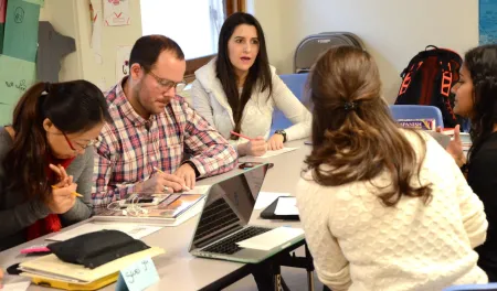 A group of five students are seated around a table having a discussion. One person is making notes on a sheet of paper.
