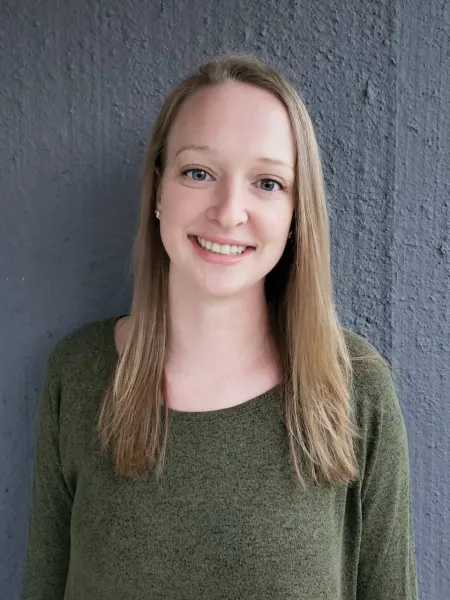 This is a photo of Courtney Yehl. Courtney is standing against a grey background. Courtney is wearing a long sleeve green shirt. 