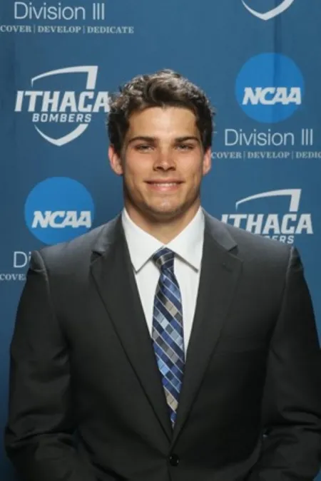 This is a photo of Jared Bauer. Jared is wearing a dark suit with a white shirt and blue striped tie. He is standing in front of a Ithaca Athletics background showing Division III athletics.