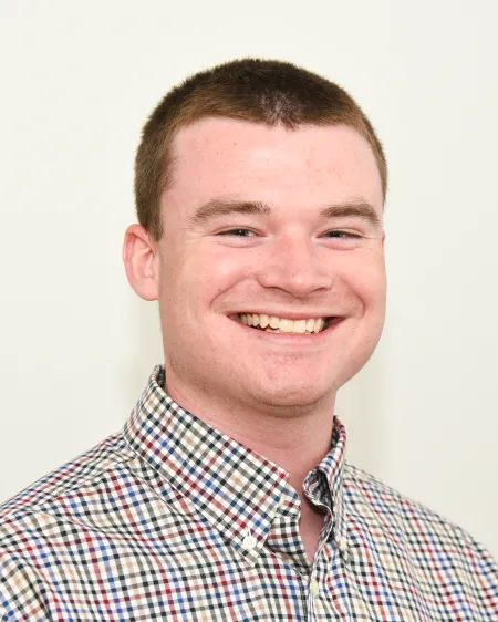 This is a photo of Jonathan Ripic. Jonathan is wearing a checkered shirt and is smiling at the camera. Jonathan has short hair, cut close.