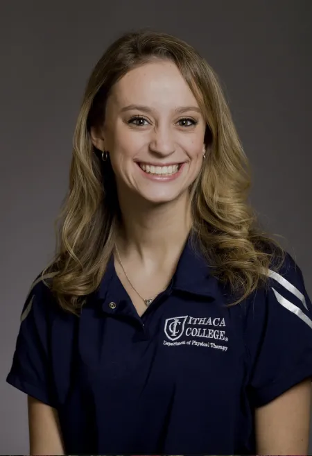This is a photo of Justina Snyder. Justina is wearing a blue Ithaca College clinic shirt and smiling at the camera.