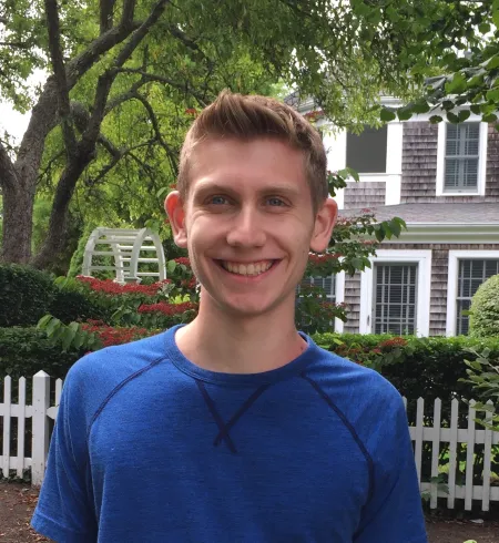 This is a photo of Matthew Darby. Matthew is wearing a long sleeve blue shirt standing in front of a white picket fence.