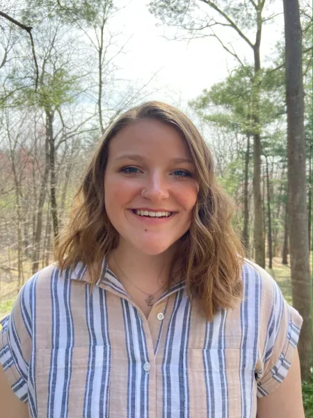 This is a photo of Michaela Zeller. Michaela is standing in the forest wearing a striped shirt with white and tan stripes.