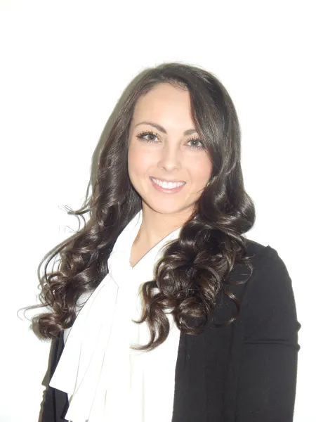 This is a photo of Natalie Knight. Natalie is wearing a white ruffled shirt underneath a black blazer. Natalie has long dark curly hair.