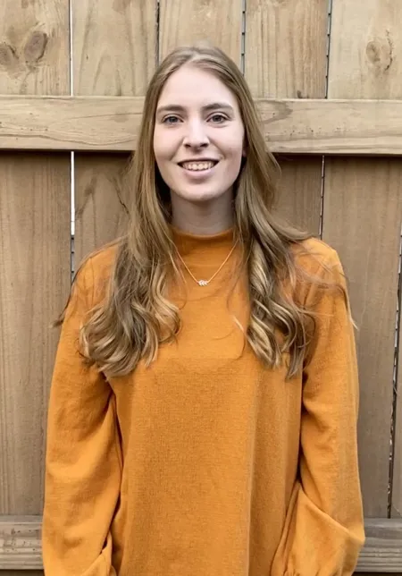 This is a photo of Paige Pickard. Paige is standing in front of a fence. Paige is wearing a long sleeve orange colored shirt.