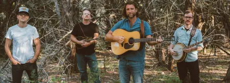 Four people standing in woods holding instruments