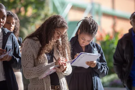 Two young women stand in an open outdoor space taking notes while listening to a speaker