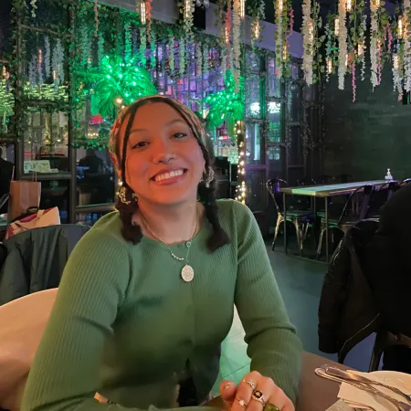 Meaghan is smiling at the camera while sitting in a room with plants behind her. She is wearing a green sweater, many rings on her fingers, and a gold necklace.