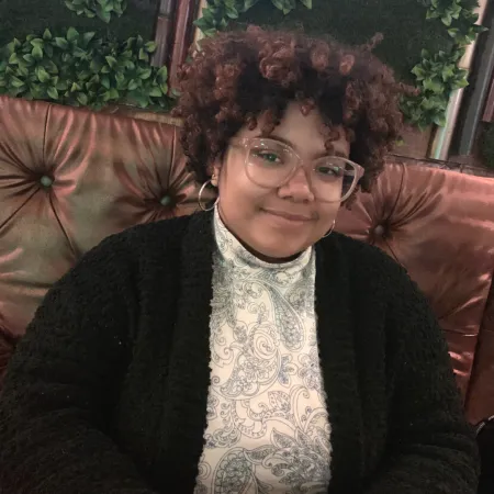 Annie is sitting in a brown upholstered booth smiling at the camera. She is wearing a black cardigan with a white blouse that has a blue paisley pattern on it. She has on blush glasses and gold hoop earrings.