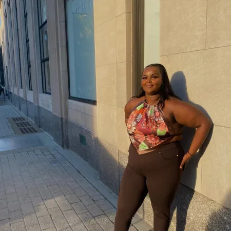 Ikraan is standing in front of a stone building. She is wearing brown pants and a multicolored blouse.