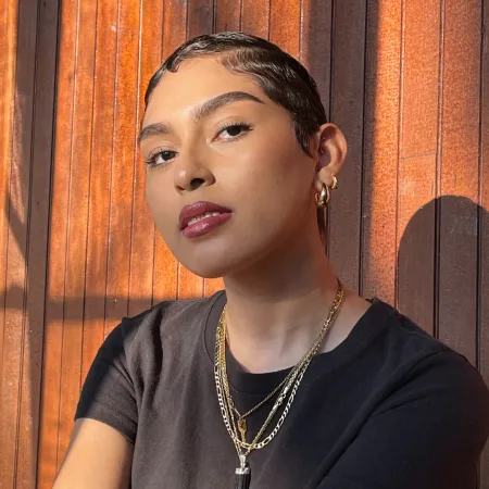 Emily is looking at the camera while wearing a black shirt and gold necklaces. She is sitting in front of a wood wall.