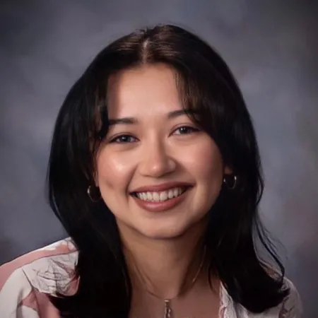 Brianna is smiling at the camera in front of a grey background. She is wearing a white and pink blouse, gold jewelry, and has her hair parted down the middle.