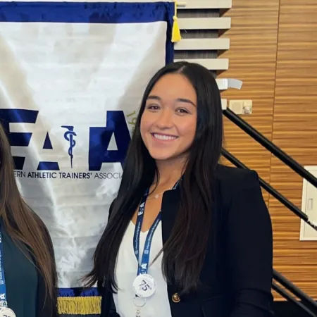 Kiana is standing in front of a white banner and a black staircase. She is smiling at the camera and wearing a black blaze with a white blouse underneath.