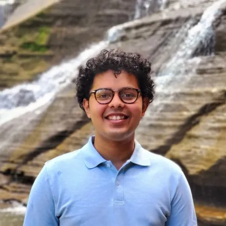 Vaibhav is smiling in front of a waterfall. He is wearing glasses and a blue polo shirt.