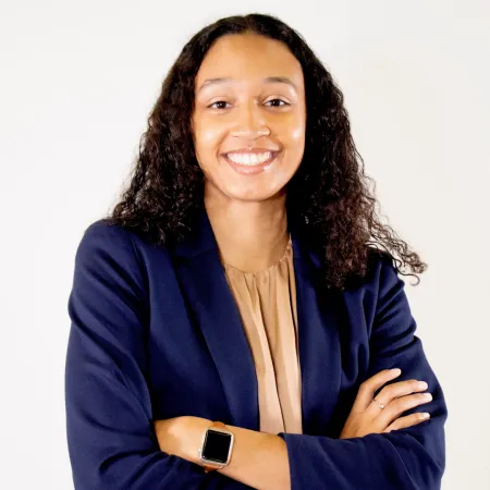 Alyssa is smiling at the camera while standing with her arms crossed in front of her body. She is wearing a blue blazer and a nude blouse.