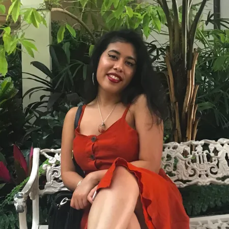 Gabrielle sits on a metal bench in front of many plants. She is wearing a red top and smiling at the camera with her hair parted to one side. 