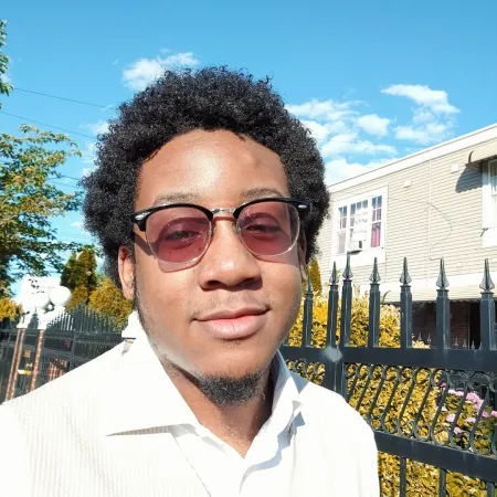 Silas is looking at the camera in front of a black fence and a blue sky. He is wearing a white polo shirt and rose tinted glasses.