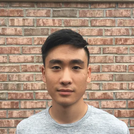 Nelson is looking at the camera in front of a brick wall wearing a grey tshirt.