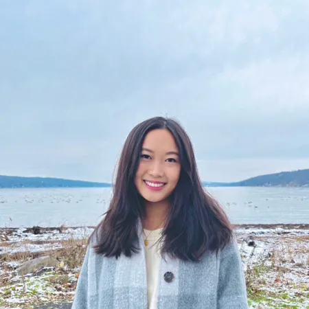 Katherine is standing in front of a short with a body of water behind her. She is wearing a large, light blue, knit sweater with a yellow shirt underneath. Her hair is parted in the middle and she is smiling at the camera.