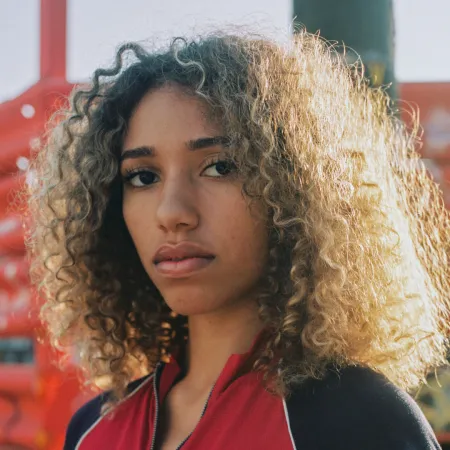 Olivia is standing and looking at the camera. She is wearing a black and red shirt. Her curly hair is parted down the middle.