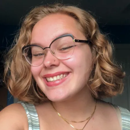 Catriona is looking at the camera and smilling. She is wearing black glasses and a gold necklace. Her hair is parted to one side and ends just above her shoulders.