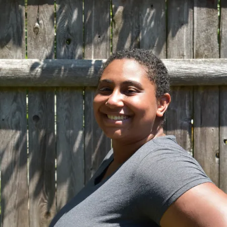 Tsedale is turned to the side, smiling at the camera in front of a wooden fence. She is wearing a grey tshirt.