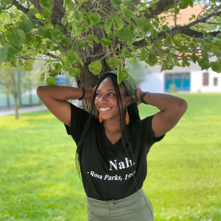 Treasure is smiling while looking off to the side while standing in front of a tree. Her hands are behind her head and she is wearing a green head scarf and a black t-shirt with the words "Nah. - Rosa Parks, 1955"