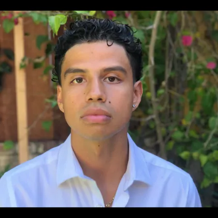 Sebastian is looking directly at the camera. He is wearing a white button up shirt and standing in front of a flowering tree.